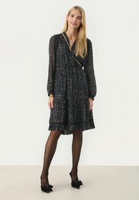 Rochie neagră cu model de puncte aurii, mâneci lungi, design înfășurat și tiv volanat. Asortată cu colanți negri transparenți și pantofi cu accent de fundiță negri.