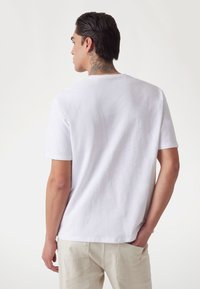 Vit t-shirt med kort ärm, tillverkad av texturerat tyg, med rund halsringning och en avslappnad passform, sedd bakifrån.