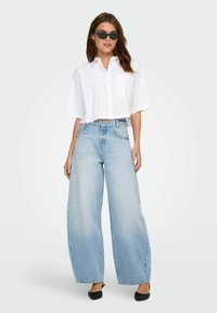 Valge lühike nööbitav särk, millel on tasku, koos helesiniste laiadest pükstest. Mudel kannab musti päikeseprille ja musti lapikuid kingi.