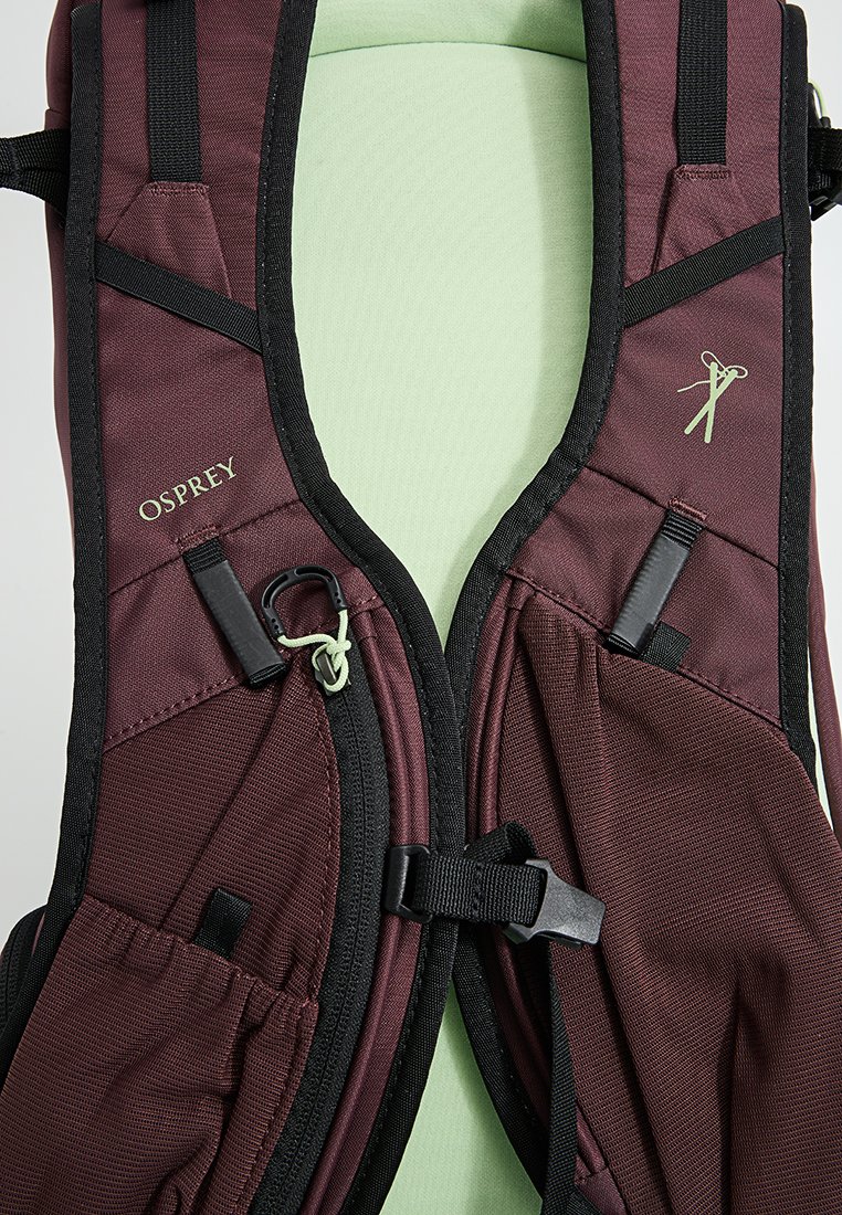 Mochila color burdeos con interior acolchado verde, que cuenta con correas ajustables, bolsillos de malla y herrajes para un cierre seguro. Logotipo de Osprey visible.