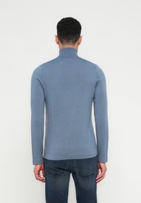 Mann in einem eng anliegenden blauen Rollkragenpullover und dunkelblauen Jeans, der vor einer schlichten weißen Wand steht.