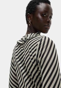 Donna con i capelli corti e ricci che indossa orecchini a cerchio dorati e una maglietta a righe bianco e nero con colletto arricciato, che guarda oltre la spalla.