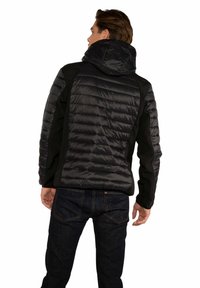 Giacca a vento nera con una texture liscia e lucida, dotata di cappuccio e pannelli laterali neri in contrasto, indossata con jeans in denim scuro.