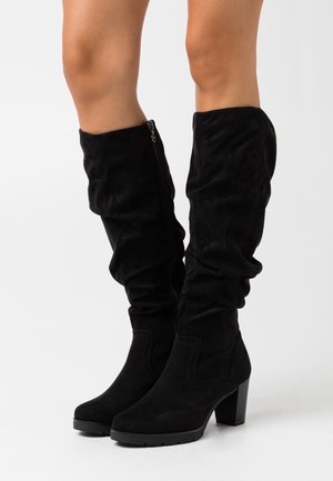 Botas negras de ante hasta la rodilla con un diseño arrugado, punta afilada y tacón grueso alto. Cremallera lateral para facilitar el uso.