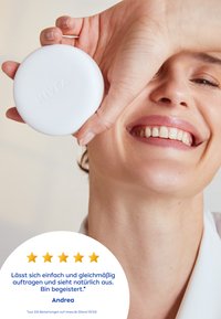 Weißer, runder Kosmetikspiegel, der in einer Hand gehalten wird, mit einem strukturierten Logo. Sanftes Licht betont einen glatten Hautton und ein natürliches Lächeln.