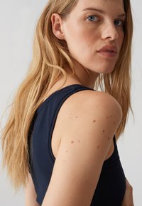 Femme aux longs cheveux blonds regarde par-dessus son épaule, portant un haut sans manches bleu marine, avec des taches de rousseur visibles sur l'épaule et le visage.