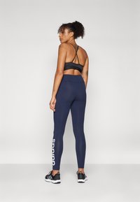 Juodas sportinis liemenėlė su juostelėmis, derinamas su tamsiai mėlynomis legging'ais su balta "adidas" logotipu. Modelis vilki juodus sportinius batus, minimalūs tekstūros ir dizaino detalės.