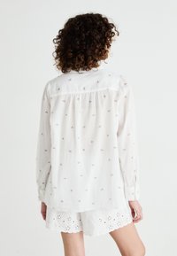 Chemise blanche à manches longues avec des broderies florales discrètes, en tissu léger, avec un dos plissé et des poignets à boutons, assortie à des shorts blancs en dentelle.