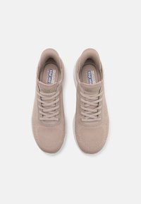 Zapatillas deportivas de color beige claro con parte superior de malla texturizada, diseño con cordones, cuello acolchado y suela de goma blanca en contraste.