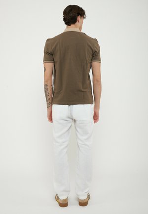 Uomo con braccio tatuato che indossa una polo marrone, pantaloni bianchi e scarpe beige, visto da dietro su uno sfondo bianco uniforme.