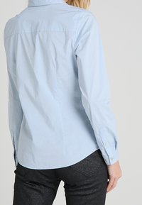 Chemise bleu clair à boutons avec un col pointu et des manches longues. Présente un ourlet arrondi et une texture de tissu lisse et uni.