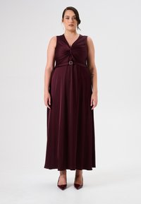 Bordeaux maxi jurk met een geknoopte voorkant, mouwloos ontwerp en een tailleband benadrukt door een zilveren gesp. Vloeiende textuur.