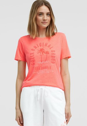 Junge Frau trägt ein korallenrotes T-Shirt mit Palmenmotiv und weiße Shorts mit Kordelzug, steht vor einem einfarbigen hellen Hintergrund.