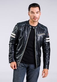 Chaqueta de cuero negra con acentos acolchados en los hombros, cremallera plateada y dos franjas blancas horizontales en las mangas. Llevada sobre una camiseta negra.