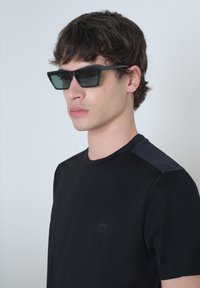 Camiseta negra con acentos texturizados en los hombros, combinada con gafas de sol negras angulares con lentes verdes. Diseño sencillo, sin patrones visibles.