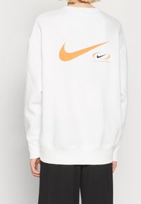 Vit sweatshirt med en stor orange Nike swoosh-logotyp på baksidan. Har ribbade ärmslut och nederkant, tillverkad av mjukt bomullsmaterial.