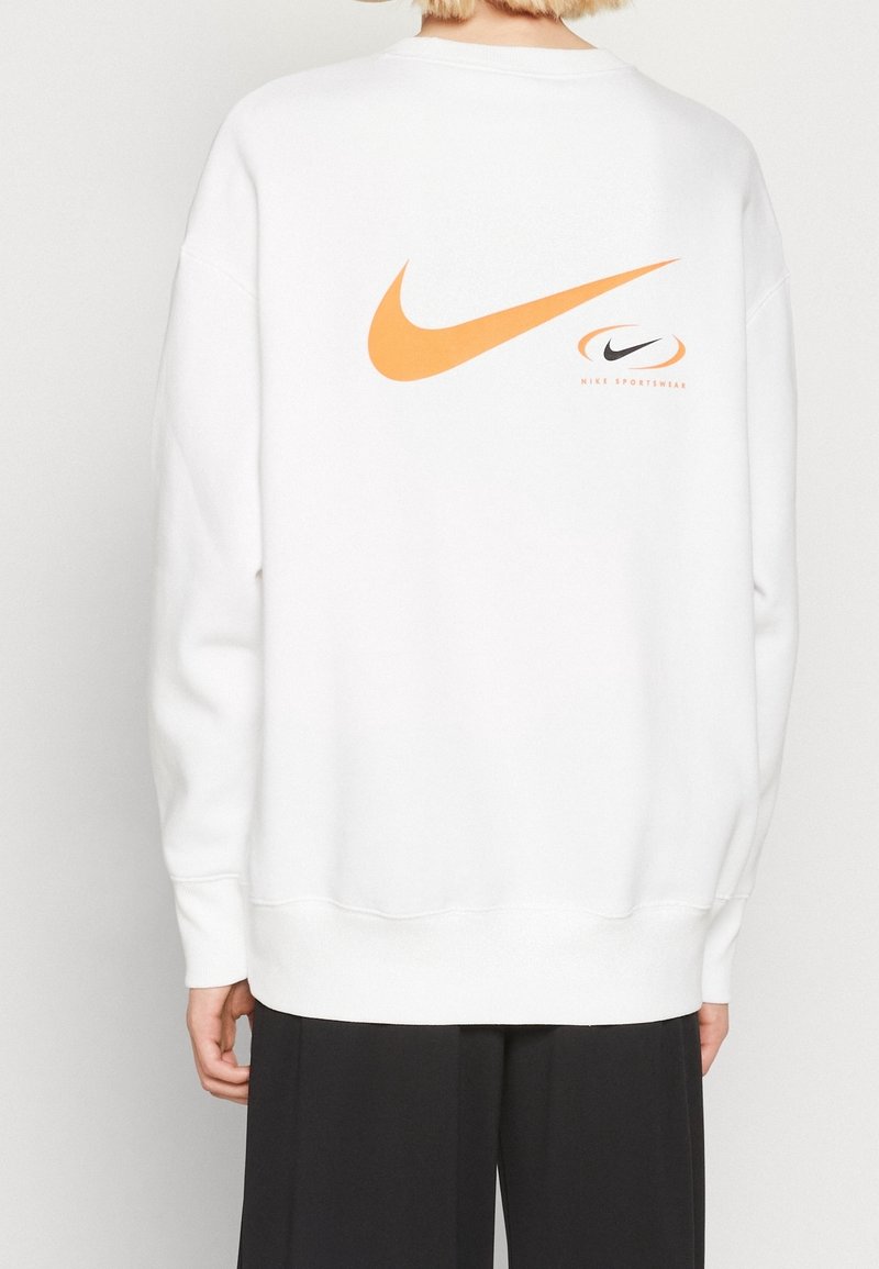 Vit sweatshirt med en stor orange Nike swoosh-logotyp på baksidan. Har ribbade ärmslut och nederkant, tillverkad av mjukt bomullsmaterial.