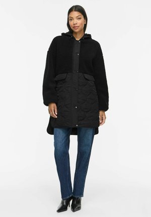 Femme portant un manteau à capuche noir avec une partie supérieure texturée et une partie inférieure matelassée, jean bleu et bottines noires, debout sur fond blanc.