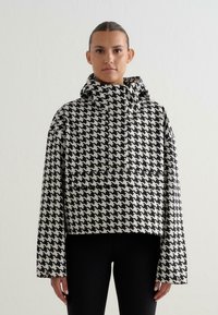 Vrouw staat rechtop, met het gezicht naar voren, gekleed in een zwart-wit houndstooth patroon met een capuchon en zwarte broek, voor een eenvoudige witte achtergrond.