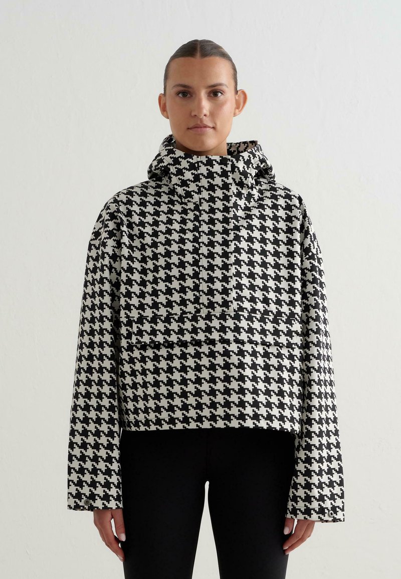 Vrouw staat rechtop, met het gezicht naar voren, gekleed in een zwart-wit houndstooth patroon met een capuchon en zwarte broek, voor een eenvoudige witte achtergrond.