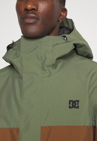 Chaqueta verde y marrón con un alto cuello, cierre de cremallera y logo en el pecho. Fabricada con una tela suave y un acabado mate.