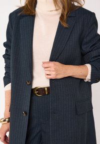 Blazer a righe blu navy realizzato in tessuto strutturato, con chiusura a pulsante singolo, abbinato a un dolcevita crema e una cintura marrone.