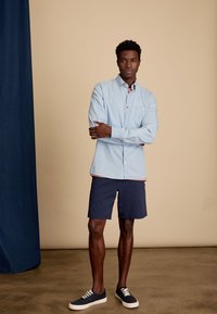Lichtblauw overhemd met knoopjes en een borstzak, marineblauwe shorts en donkere sneakers. Eenvoudig ontwerp, casual pasvorm, gladde textuur.