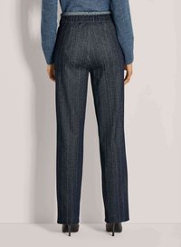 Vrouw in hoge taille, donkergrijze pinstripe broek en zwarte hoge hakken, staand tegen een effen lichte achtergrond.