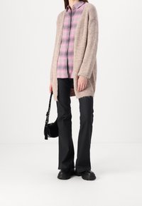 Beżowy dziergany kardigan na różowej koszuli w kratę zapinanej na guziki, w połączeniu z czarnymi rozszerzanymi spodniami i czarnymi masywnymi butami. Czarna torebka na ramię.