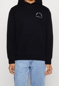 Černá mikina s kapucí a přední kapsou, na hrudi s malým bílým logem "Tommy Hilfiger New York", nošena se světle modrými džíny.