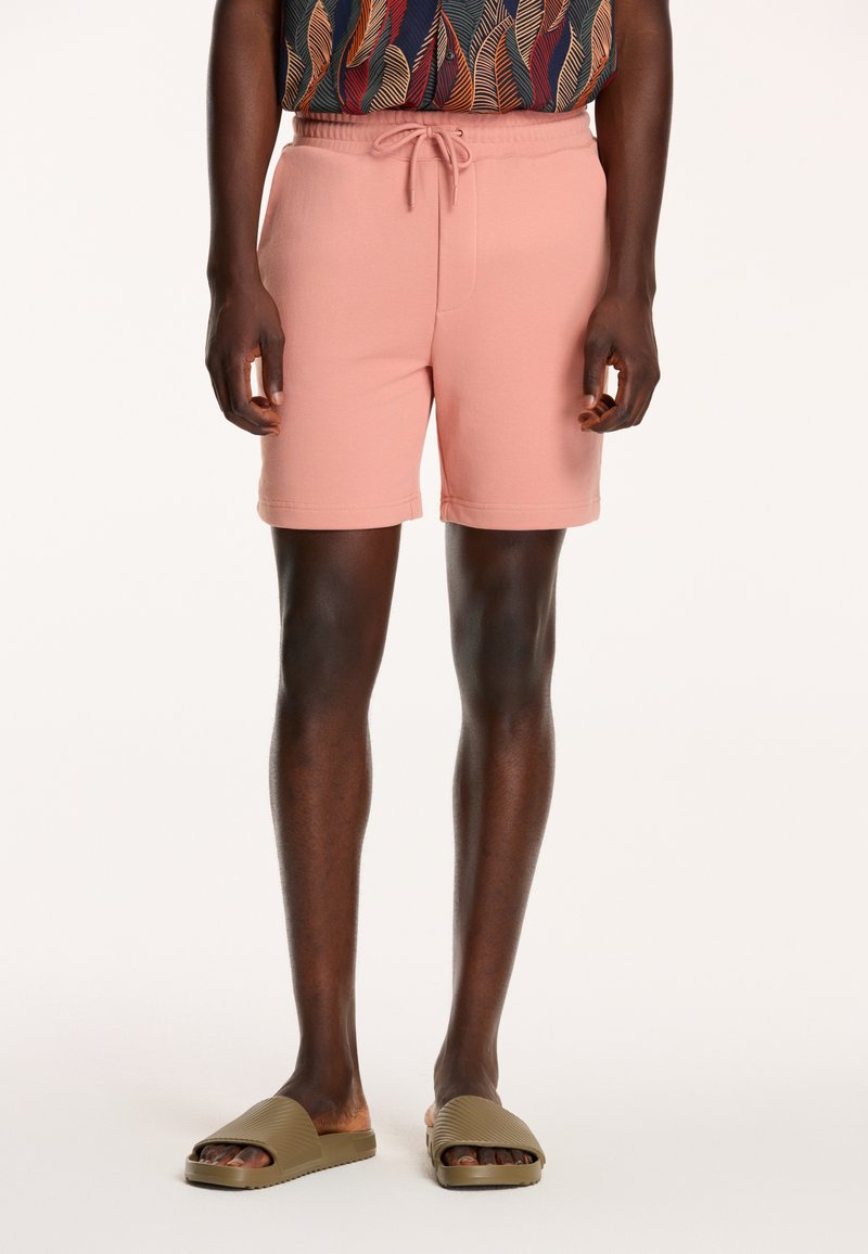 Herrshorts i persikofärg med elastisk midjeband och snörning; mjuk textur; tillsammans med olivgröna sandaler.