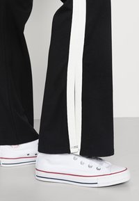 Calças pretas com riscas laterais brancas, combinadas com ténis clássicos de lona brancos com acentos vermelhos e azuis. Sapatos baixos com ilhós de metal.