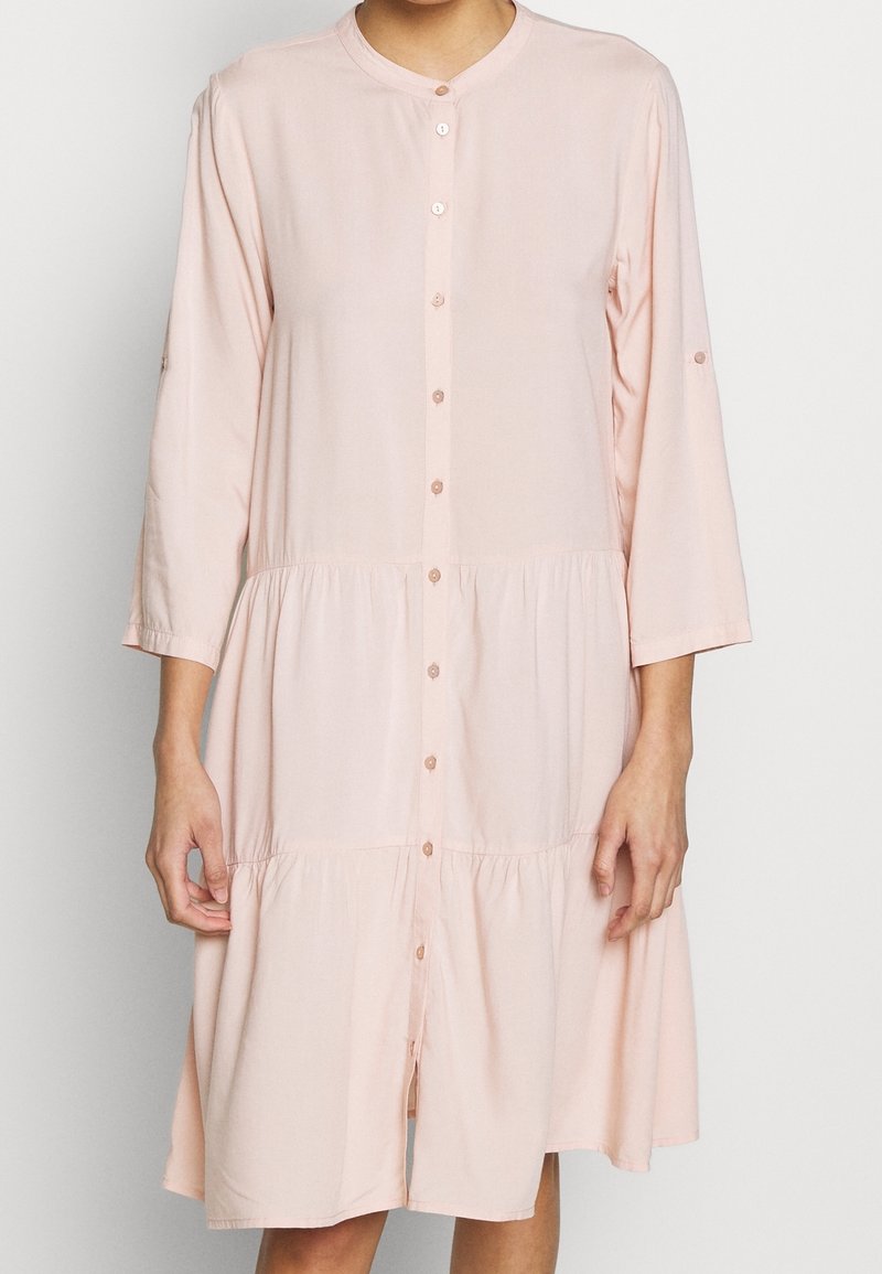 Abito rosa chiaro a balze, con bottoni e colletto rotondo, maniche a tre quarti. Tessuto morbido con una texture liscia, design minimalista.