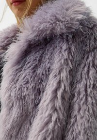 Pelsfrakke i blød lavendel, der har en lang, shaggy tekstur med en bred krave, der tilfører volumen og varme. Bemærkelsesværdig for sit bløde udseende.