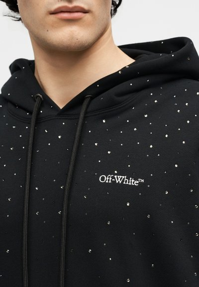 Tuvplāns ar personu, kas valkā melnu Off-White kapuci, uz kura priekšpuses redzamas nelielas atstarojošas punktiņas un aukliņas.