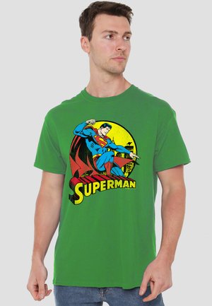 Young man facing right wearing green T-shirt with vintage Superman graphic and yellow city skyline background.