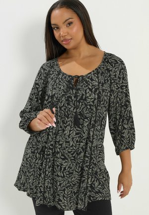Frau trägt schwarze Tunika mit beigem Blattmuster und Dreiviertelärmeln, posiert vor einfachem weißem Hintergrund.