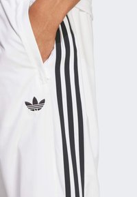 Fehér sportmelegítő nadrág három fekete oldalsávval, cipzáras zsebekkel és hímzett fekete Adidas logóval a combon.