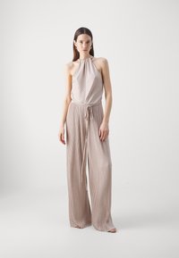Woman wearing sleeveless, pleated beige jumpsuit with halter neck and wide-leg pants, standing against a plain white background.