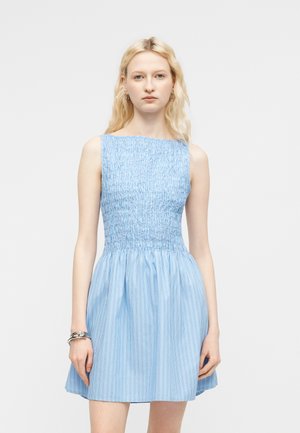 Γυναίκα με μακριά ξανθά μαλλιά, φορώντας αμάνικο ανοιχτό μπλε φόρεμα με ρεγκλάν κορσέ και λεπτές κάθετες ρίγες, στέκεται μπροστά σε απλό φόντο.