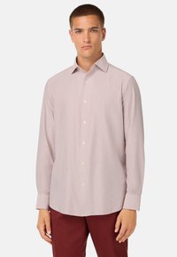 Una camicia a maniche lunghe di colore rosa chiaro con un sottile motivo a righe, colletto classico e orlo arrotondato, abbinata a pantaloni bordeaux scuri.