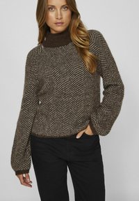 Maglione marrone con motivo a spina di pesce, colletto alto e maniche larghe. Realizzato in un tessuto a maglia testurizzato. Vengono indossati pantaloni neri.