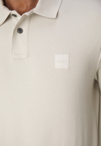 Lys beige polo shirt med en tekstureret fabric, to knapper og et syet logo patch i et kvadrat på venstre bryst.