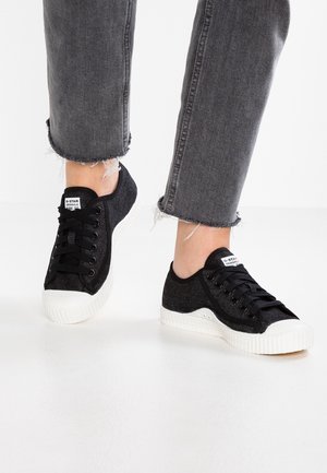 Zwarte canvas sneakers met witte rubberen neuskapjes en gestructureerde bovenzijde, gecombineerd met versleten grijze spijkerbroeken.