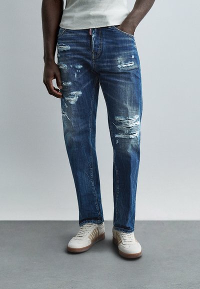 Homme portant un jean bleu déchiré et des baskets blanches avec des rayures beiges, debout avec une main dans la poche contre un fond uni.