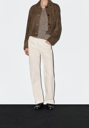 Persona che indossa una giacca in camoscio marrone, un maglione grigio lavorato a maglia, pantaloni bianchi a gamba dritta e mocassini neri lucidi, in piedi con una mano in tasca.