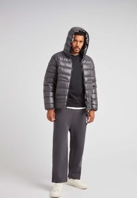 Chaqueta acolchada gris con capucha, cierre frontal y detalles de logo. Lleva puesta una sudadera negra y pantalones de chándal grises, con zapatillas blancas.
