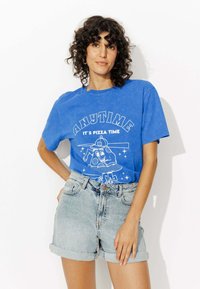 Femme aux cheveux bouclés portant un T-shirt bleu avec l'inscription "TOUJOURS LE TEMPS POUR LA PIZZA" et un short en denim clair, posant contre un fond blanc.