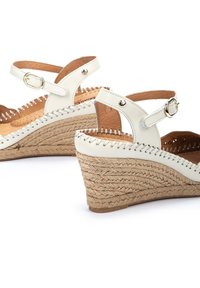 Sandalias de cuña de cuero blanco con suelas de yute trenzado, que presentan una correa de tobillo ajustable, costuras decorativas y acentos metálicos redondos.