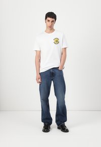 T-shirt en coton blanc avec un imprimé graphique jaune et noir portant les mots "SUNNIER POOLBAR". Associé à un jeans en denim bleu et des chaussures noires.